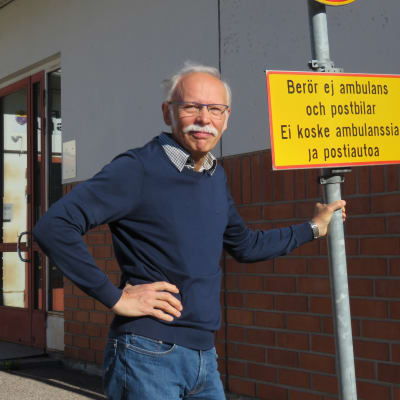 En man med grått hår, mustasch och blå tröja står utanför Ingå hälsocentrals ingång. Han står och håller i en trafikskylt som förbjuder trafik, men med tillägget "berör ej ambulans och postbil".