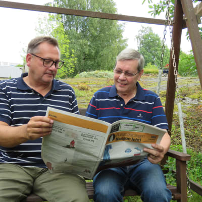 Två män sitter i en trädgårdsgunga och tittar i en papperstidning, Hangötidningen. Sommar, utomhus.
