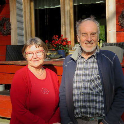En äldre kvinna och äldre man står framför en rödmålad stockstuga. De ser in i kameran och ler.