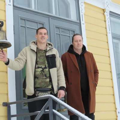 Porträttbild på Nico Rönnberg och Andreas Rönnberg utanför Karis stationshus, ett gult trähus. Nico håller i en stationsklocka som hänger från husväggen. Vinter och snö.