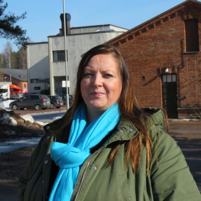 Profilbild på Piia Hasselberg, hon står utomhus, mars 2022, bakom röd tegelbyggnad på gamla Ekåsenområdet.