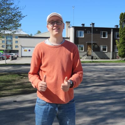 En ung man med beigefärgad keps, skärmmössa och orangefärgad tröja står och håller upp tummarna. Han ser glad ut.