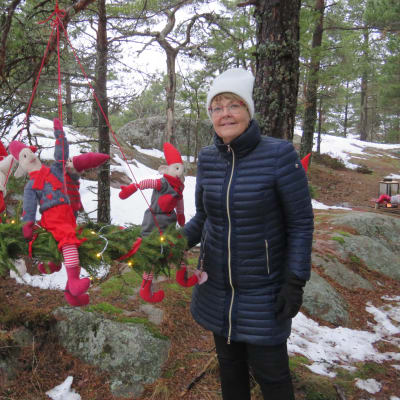 En kvinna i vinterkläder står bredvik en juldekoration hon gjort. Det är en grankrans som hänger från en trädgren med hjälp av röda band. I kransen sitter röda tygtomtar som kvinna gjort
