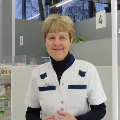 En kvinna i vit arbetsrock i ett apotek. Hon står framför betjäningsdiksen i Ingå apotek, ser in i kameran och ler.