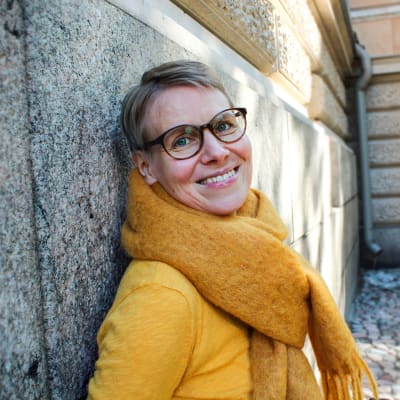 En yngre brett leende, glasögonprydd kvinna klädd i gul tröja och gul halsduk lutar sig mot en husgrund 