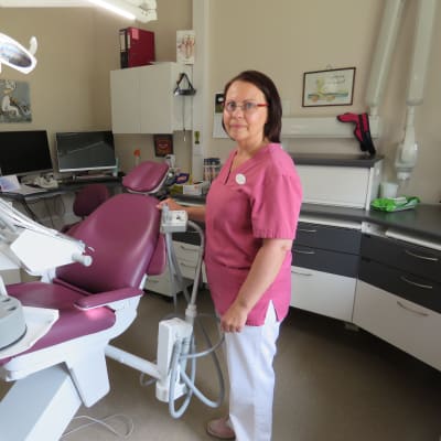 En kvinna, tandläkare, i rosa arbetsrock och vita byxor står vid en rosa tandläkarstol.