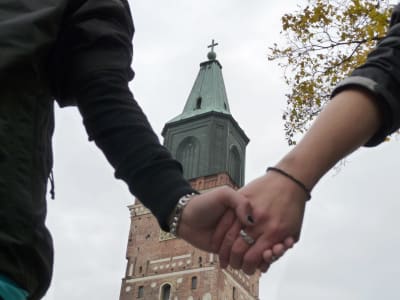 Två kvinnohänder med förlovningsringar håller i varandra med Åbo domkyrka i bakgrunden.