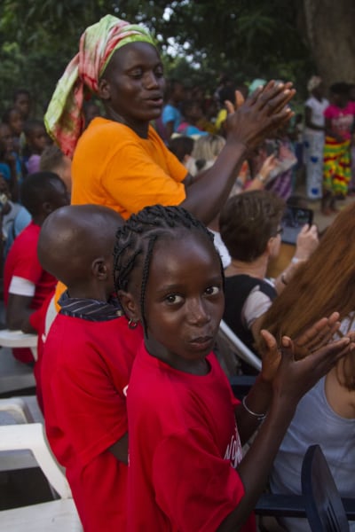 Suuri joukko käsiään taputtavia ihmisiä afrikkalaisessa kylässä, etualalla kaksi punaiseen t-paitaan pukeutunutta nuorta tyttöä, joista etummainen katsoo kameraan.