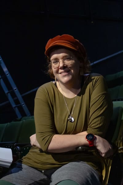 På bilden ses en teaterregissör iklädd en orange keps och grön tröja. Hon sitter i en teatersalong.