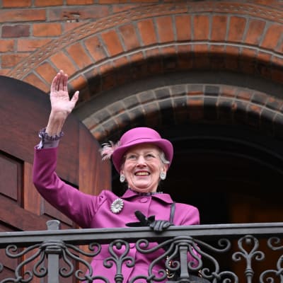 Drottning Margrethe iklädd lila hatt och kappa vinkar glatt på en balkong.