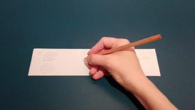 En hand håller i en penna vid en vit papperslapp. 