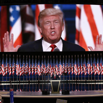 Donald Trump står bakom ett guld- och svartfärgat podium med flera amerikasnka flaggor och en enorm screen bakom honom som streamar talet den 21 juli 2016.