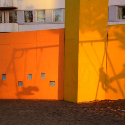 En orange husvägg med skuggorna från barn som gungar och vuxna som ger fart i en lekpark.