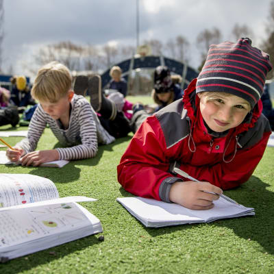 Elever i S:t Karins svenskspråkiga skola ligger på en gräsmatta och skriver i häften.