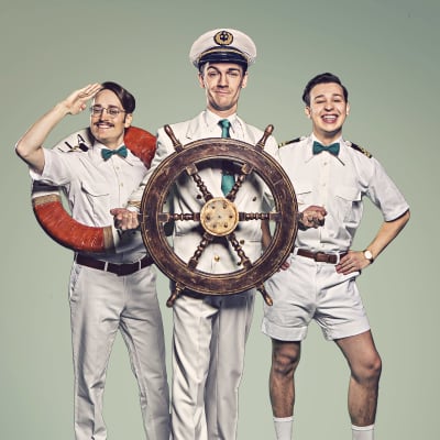 Tre unga män i sjömanskostymer poserar för kameran. Mannen i mitten har ett roder i handen. Mannen till vänster har en livboj runt halsen