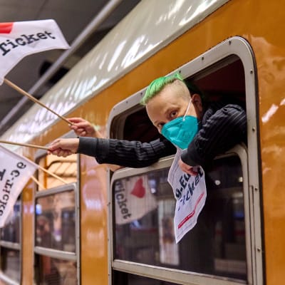 En grönhårig tysk resenär hänger ut från fönstret på ett orangefärgat tåg med en flagga med texten "jag älskar nio euros biljetten" i handen. 