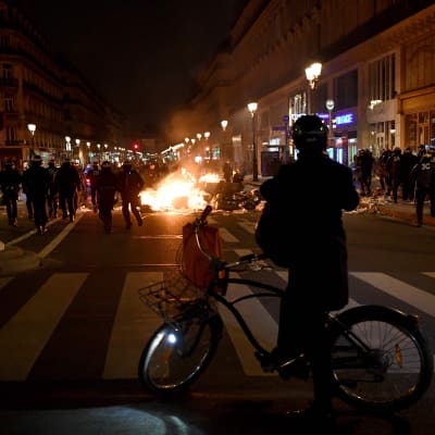 Pyöräilijä seisoo kuvan etuosassa katsomassa taustalla palavaa mielenosoittajien sytyttämää paloa.