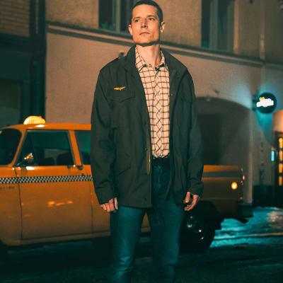 Mies (näyttelijä Max Ovaska) seisoo maiharitakissa öisellä kadulla amerikkalaismallisen keltaisen taksin vieressä. Mukaelma elokuvasta Taksikuski.