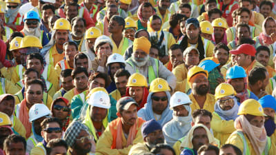 Suuri joukko miespuolisia työntekijöitä istuu ulkona. Monilla on päässä keltainen rakennustyöläisen kypärä. 