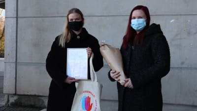 En ljushårig kvinna och en mörkhårig kvinna, som båda har munskydd på sig, håller upp ett diplom och en blombukett som de fått av Röda Korset.