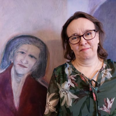 Ett porträtt av en lilaklädd, gråhårig kvinna och på hennes högra sida en verklig kvinna, leende, brunhårig, glasögonförsedd och klädd i mönstrad blus.