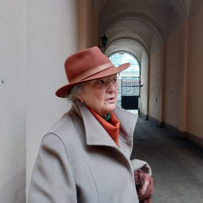 En mycket elegant dam i grå kappa, bruna läderhandskar, orange scarf och aningen ljusarebrun filthatt står i ett portvalv och pratar fundersamt.