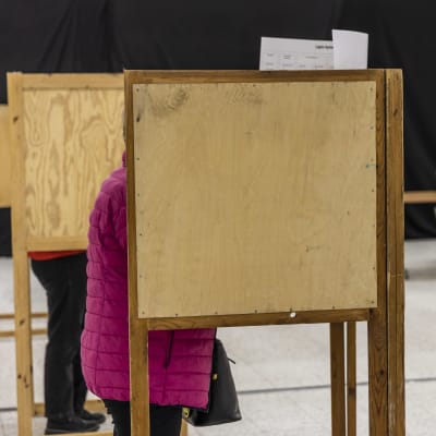 I förgrunden kvinna i rosa ytterock som står i valbås för att avlägga sin röst. I bakgrunden valfunktionär som stämplar valsedel.