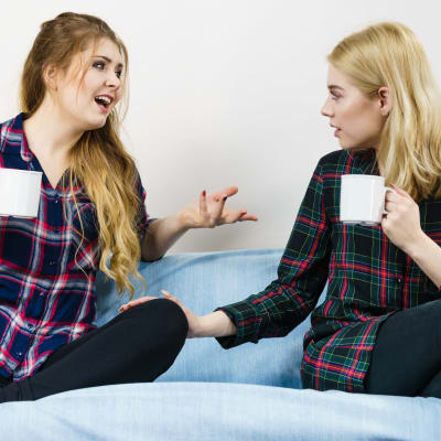 Två kvinnor med kaffekoppar i soffa, den ena gestikulerar och pratar, den andra sitter tyst