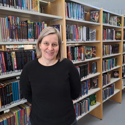 En leende kvinna med blont hår i pagefrisyr framför bokhyllor på ett bibliotek.