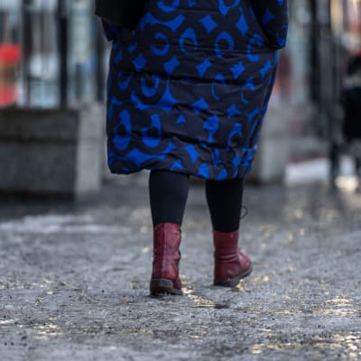 En person i blå jacka går på en trottoar. Den är täckt av ett lager is med sand på.
