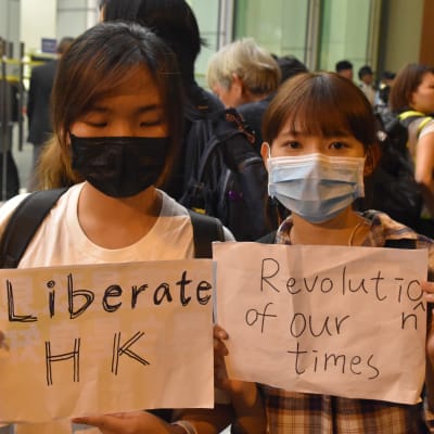 Två kvinnor i munskydd håller upp skyltar där det på engelska står "berfria Hongkong" och "vår tids revolution"