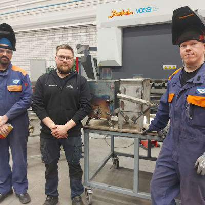 Kolme miestä seisoo työvaatteissa hitsauspäydän edessä. Pöydällä on kamiina.