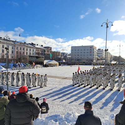 Solen lyser över tiotals beväringar som deltar i försvarsmaktens paradmönstring på Vasa torg. I förgrunden publik som samlats. 