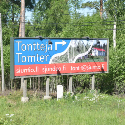 en skylt längs en väg där det står tomter till salu på svenska och finska och sedan sjundeå.fi, siuntio.fi