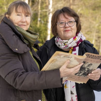 Två kvinnor står utomhus och håller i en gammal tidning som heter Kvinnotid / Aikanainen.