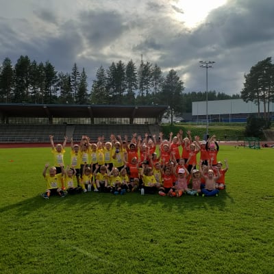 Keltapaitaiset pojat ja oranssipaitaiset tytöt nostavat käsiä ilmaan jalkapallokentällä.