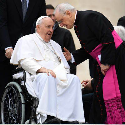 Påve Franciskus iklädd vitt sitter i en rullstol omgiven av svartklädda kardinaler.