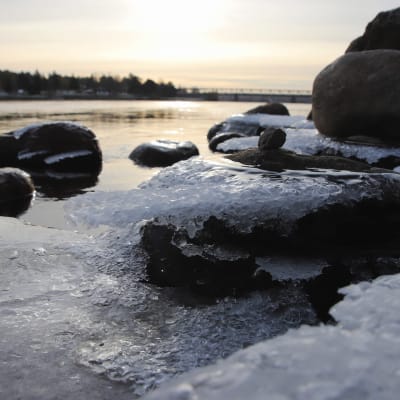 Joen kivikkoista rantaa syksyisessä auringonpaisteessa, kiviin on muodostunut jäätä.