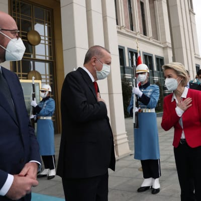 Två kostymklädda män och en kvinna i röd jacka och svarta byxor står utanför en byggnad med två soldater med gevär och långa ljusblå vapenrockar bakom sig. Alla bär munskydd. Från vänster Charles Michel, Recep Tayyip Erdogan och Ursula von der Leyen.