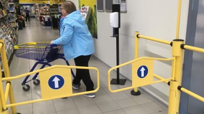 Nainen työntää ostoskärryjä kaupan portilla, jossa on käsidesiautomaatti. 