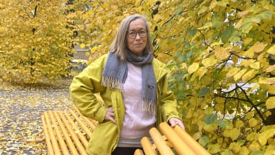 Leena Stolzmann har på sig en gul jacka och står omgiven av gula höstlöv. Hon ser allvarlig ut.