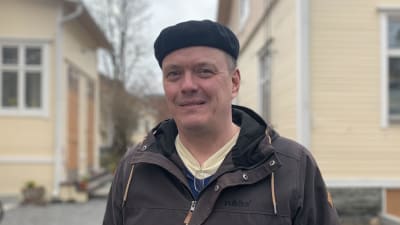Mansperson står intill träshusfasad, blicken riktad in i kameran.