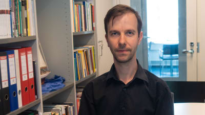 Johannes Kananen i en svart skjorta stående bredvid en bokhylla.