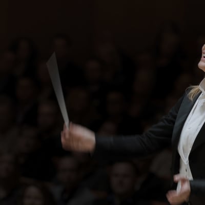 En kvinnlig dirigent höjer taktpinnen medan hon lutar sig bakåt.