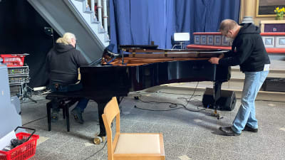 Två män håller på och gör något med sladdar och ett piano.