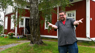 En man i kortärmad skjorta sträcker fram händerna i en "Spider man"-gest. I bakgrunden en rödmålad bondstuga.