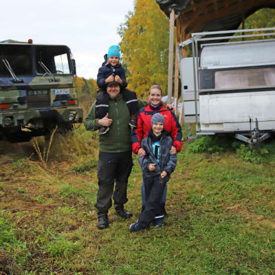 Nissisen nelihenkinen perhe, taustalla maastokuorma-auto ja asuntovaunu.