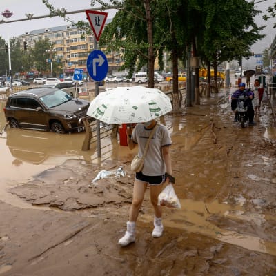 En ung kvinna går med ett paraply och en plastkasse över en lertäckt gata i Beijing.