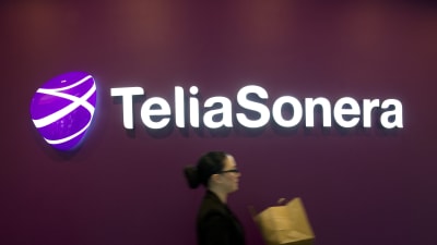 Telia Soneras logotyp mot en vinröd bakgrund. I förgrunden suddigt en kvinna som går förbi.