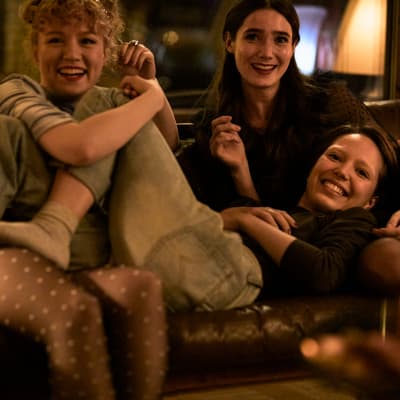 Kuvassa kolme tyttöä istuu sohvalla sylikkäin ja nauraa.
